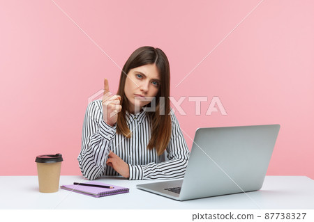 Be careful. Bossy strict business woman in striped shirt warning with admonishing finger gesture, giving advice while working on laptop at home office. Indoor studio shot isolated on pink background Be careful. Bossy strict business woman in striped shirt warning with admonishing finger gesture, giving advice while working on laptop at home office. Indoor studio shot isolated on pink background 87738327