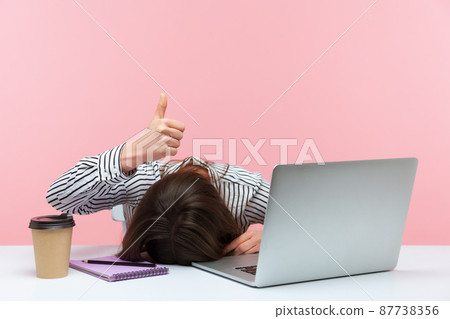 I am fine. Extremely tired sleepy woman lying on table with laptop and cup of coffee showing thumbs up, exhausted with overwork, vitamin deficiency. Indoor studio shot isolated on pink background 87738356