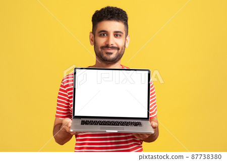 Positive cheerful man with beard in striped t-shirt holding white display laptop and looking at camera with smile, satisfied with new operating system. Indoor studio shot isolated on yellow background 87738380