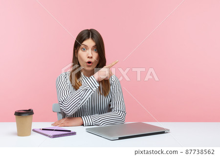 Excited shocked woman pointing finger away paying your attention at empty space for advertising banner, looking at camera with astonishment. Indoor studio shot isolated on pink background 87738562