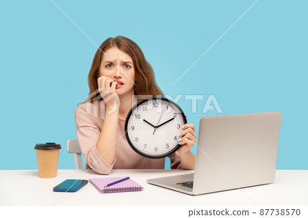 Upset anxious woman, office employee sitting at workplace, holding big clock and biting nails, working late hours, worrying about deadline, overtime job. indoor studio shot isolated on blue background Upset anxious woman, office employee sitting at workplace, holding big clock and biting nails, working late hours, worrying about deadline, overtime job. indoor studio shot isolated on blue background 87738570
