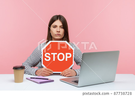 Serious woman office worker holding stop symbol, warning with red traffic sign and looking angrily, sitting at workplace with laptop, home office. Indoor studio shot isolated on pink background 87738728