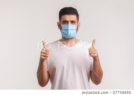 Effective protection against contagious disease. Man showing thumbs up and wearing hygienic mask to prevent infection, respiratory illness such as flu, coronavirus. indoor studio shot, isolated Effective protection against contagious disease. Man showing thumbs up and wearing hygienic mask to prevent infection, respiratory illness such as flu, coronavirus. indoor studio shot, isolated 87738848