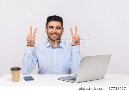 This is win. Joyful businessman sitting office workplace with laptop on desk, showing victory gesture, celebrating successful project, job promotion. indoor studio shot isolated on white background This is win. Joyful businessman sitting office workplace with laptop on desk, showing victory gesture, celebrating successful project, job promotion. indoor studio shot isolated on white background 87738917