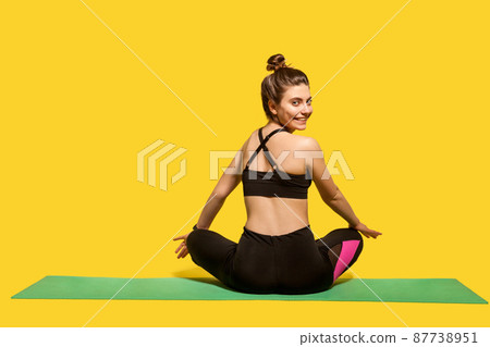 Back view cheerful woman with hair bun in tight sportswear sitting in lotus position on gym mat practicing yoga, turning head to camera and smiling joyfully, doing sports. Happy healthy lifestyle 87738951