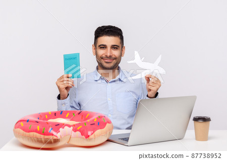 Cheerful man holding paper airplane and passport in hands, sitting in office workplace with rubber ring on desk, planning travel vacation, weekend trip. studio shot isolated on white background 87738952