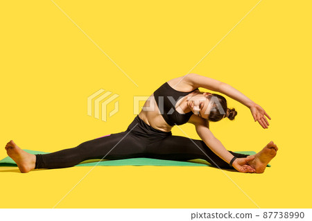 Sporty gymnast woman with hair bun in tight sportswear sitting on mat with spread legs, bending to touch toe, stretching back and hands muscles, flexibility exercises. studio shot, isolated on yellow Sporty gymnast woman with hair bun in tight sportswear sitting on mat with spread legs, bending to touch toe, stretching back and hands muscles, flexibility exercises. studio shot, isolated on yellow 87738990