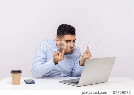 Annoyed angry businessman sitting office workplace, showing middle finger to laptop screen, aggressively communicating on video call, expressing hate to job or coworker. indoor studio shot isolated 87739075