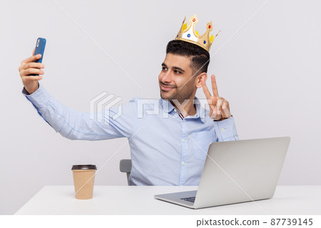 Proud happy businessman sitting in office workplace, wearing crown on head and taking selfie on mobile phone, showing his success in social networks. indoor studio shot isolated on white background 87739145