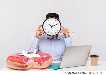 Time to rest. Unknown man hiding face behind clock, sitting in office workplace with rubber ring passport and paper airplane on desk, employee asking for break, relax and travel vacation. studio shot 87739199