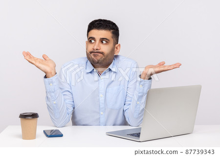 Dont know. Puzzled clueless confused businessman sitting office workplace with laptop on desk, shrugging shoulders, doubting cant make decision. indoor studio shot isolated on white background Dont know. Puzzled clueless confused businessman sitting office workplace with laptop on desk, shrugging shoulders, doubting cant make decision. indoor studio shot isolated on white background 87739343