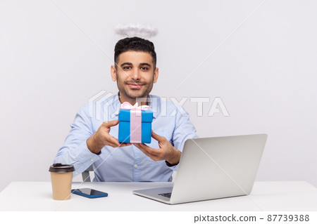 Congratulations. Happy angelic businessman with nimbus on head holding gift box, sitting at laptop workplace, greeting with professional holidays. indoor studio shot isolated on white background Congratulations. Happy angelic businessman with nimbus on head holding gift box, sitting at laptop workplace, greeting with professional holidays. indoor studio shot isolated on white background 87739388