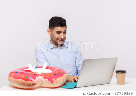 Man booking tickets or tour online using laptop, looking shocked surprised by price, searching low cost, sitting surrounded by rubber ring passport and paper airplane on desk. studio shot isolated Man booking tickets or tour online using laptop, looking shocked surprised by price, searching low cost, sitting surrounded by rubber ring passport and paper airplane on desk. studio shot isolated 87739389