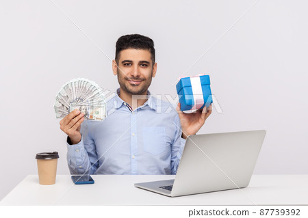 Purchase of holiday gifts, internet shopping. Happy elegant man sitting workplace with laptop, holding money dollars and present box, smiling at camera. indoor studio shot isolated on white background 87739392