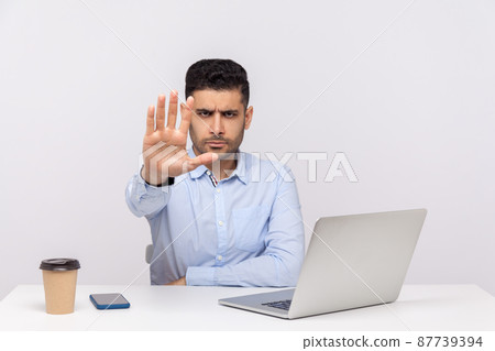 Strict bossy elegant young businessman sitting in office workplace with laptop and coffee, warning with stop gesture, denial, prohibition hand sign. indoor studio shot isolated on white background 87739394