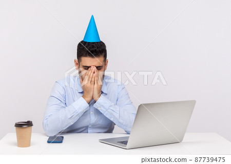 Upset depressed employee guy with party cone on head crying at workplace, hiding face feeling desperate lonely on birthday has to stay in office. indoor studio shot isolated on white background Upset depressed employee guy with party cone on head crying at workplace, hiding face feeling desperate lonely on birthday has to stay in office. indoor studio shot isolated on white background 87739475
