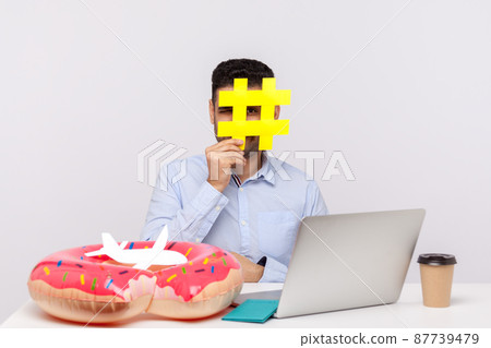 Time to rest. Man looking through hashtag symbol, sitting in office workplace with rubber ring passport and paper airplane on desk, holding hash sign of viral web topic about relax travel. studio shot 87739479