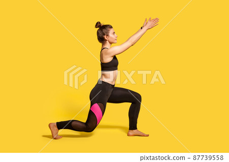Side view, slim young woman with hair bun in tight sportswear doing sport lunge exercise, standing one knee and raising hands, warming up training muscles. full length studio shot, isolated on yellow 87739558