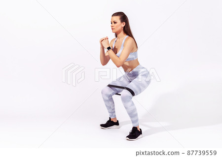 Full length side view assertive woman gymnast in white sportswear doing squats with resistance band on knees, pumping legs and glutes muscles. Indoor studio shot isolated on gray background 87739559
