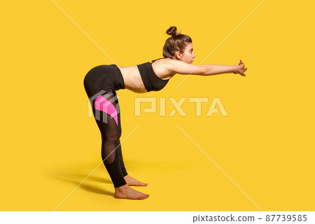 Side view, athletic woman with hair bun in tight sportswear doing sport, bending and stretching hands out, warming up training muscles for flexibility. full length studio shot, isolated on yellow 87739585