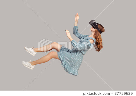 Gamer girl in ruffle dress hovering in air, levitating with virtual reality glasses on head, playing game through vr headset, floating in cyberspace. indoor studio shot isolated on gray background 87739629
