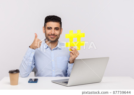 Businessman sitting in office workplace with laptop, holding big hashtag symbol and showing thumbs up, like gesture, hash sign of viral internet idea, web seo promotion. indoor studio shot isolated Businessman sitting in office workplace with laptop, holding big hashtag symbol and showing thumbs up, like gesture, hash sign of viral internet idea, web seo promotion. indoor studio shot isolated 87739644