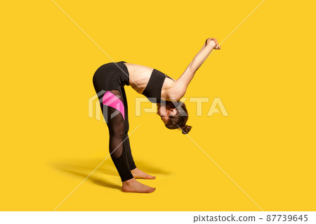 Athletic girl with hair bun in tight sportswear doing sport, bending and stretching hands backwards, warming up training muscles for flexibility. full length studio shot, isolated on yellow background Athletic girl with hair bun in tight sportswear doing sport, bending and stretching hands backwards, warming up training muscles for flexibility. full length studio shot, isolated on yellow background 87739645