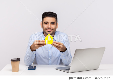 Portrait of real estate agent man sitting in office workplace and holding paper house, smiling friendly, advertisement of purchase or rental housing. indoor studio shot isolated on white background Portrait of real estate agent man sitting in office workplace and holding paper house, smiling friendly, advertisement of purchase or rental housing. indoor studio shot isolated on white background 87739646