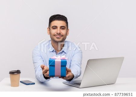 Positive elegant businessman sitting in office workplace, giving gift box to camera and smiling friendly, holding present bonus, congratulating on professional holiday. indoor studio shot isolated 87739651