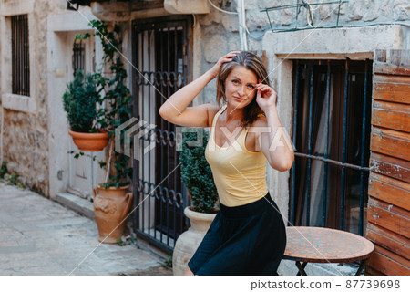 Girl Tourist Walking Through Ancient Narrow Street On A Beautiful Summer Day In MEDITERRANEAN MEDIEVAL CITY, OLD TOWN BUDVA, MONTENEGRO. Young Beautiful Cheerful Woman Walking On Old Street At Girl Tourist Walking Through Ancient Narrow Street On A Beautiful Summer Day In MEDITERRANEAN MEDIEVAL CITY, OLD TOWN BUDVA, MONTENEGRO. Young Beautiful Cheerful Woman Walking On Old Street At 87739698