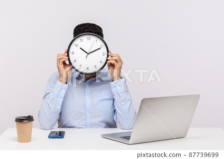 Time to work. Male employee sitting in office workplace and covering face with big clock, meeting deadline, time management concept, business schedule. indoor studio shot isolated on white background Time to work. Male employee sitting in office workplace and covering face with big clock, meeting deadline, time management concept, business schedule. indoor studio shot isolated on white background 87739699