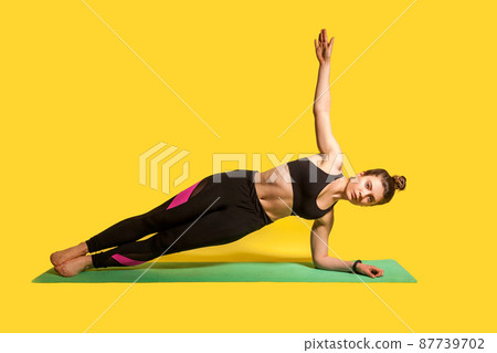 Side plank pose. Young fitness woman in tight sportswear practicing yoga, doing Vasisthasana exercise with one hand raised, training muscles. studio shot, sport workouts isolated on yellow background Side plank pose. Young fitness woman in tight sportswear practicing yoga, doing Vasisthasana exercise with one hand raised, training muscles. studio shot, sport workouts isolated on yellow background 87739702