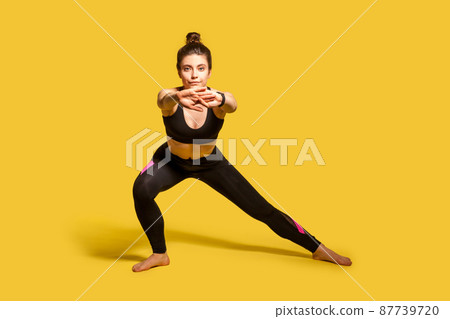 Athletic pretty girl with hair bun in tight sportswear doing sport, lower-body exercise, stretching hands out, warming up training muscles for flexibility. full length studio shot, isolated on yellow Athletic pretty girl with hair bun in tight sportswear doing sport, lower-body exercise, stretching hands out, warming up training muscles for flexibility. full length studio shot, isolated on yellow 87739720