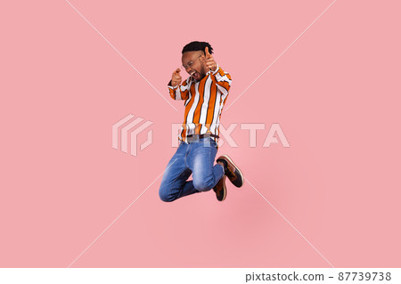 Full length happy successful african man with dreadlocks in striped shirt denim pants actively jumping pointing fingers at camera, celebrating victory. Indoor studio shot isolated on pink background Full length happy successful african man with dreadlocks in striped shirt denim pants actively jumping pointing fingers at camera, celebrating victory. Indoor studio shot isolated on pink background 87739738
