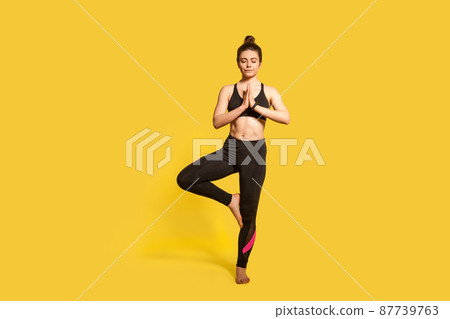 Tree pose. Athletic slim woman with hair bun in tight sportswear practicing yoga, doing Vrksasana exercise on one leg, hands in prayer namaste gesture. full length studio shot, sport workouts isolated 87739763