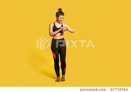 Cheerful athletic woman with hair bun in tight sportswear touching wristwatch display, checking pulse time on fitness tracker, smiling satisfied with health indicators after trainings. Sport activity 87739764