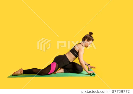 Sportswoman unfolding mat, starting day with workout, practicing yoga, one legged king pigeon pose, doing pilates exercise, warming up with flexibility exercises. studio shot, yellow background Sportswoman unfolding mat, starting day with workout, practicing yoga, one legged king pigeon pose, doing pilates exercise, warming up with flexibility exercises. studio shot, yellow background 87739772