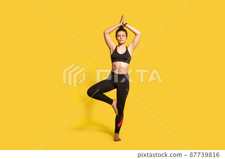 Tree pose. Athletic concentrated woman in tight sportswear practicing yoga, meditating with closed eyes, doing Vrksasana exercise on one leg, hands up. full length studio shot, sport workouts isolated 87739816