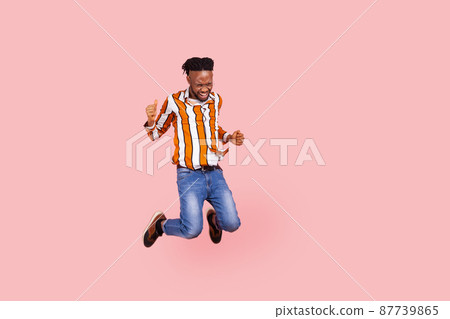 Full length happy positive african man with dreadlocks in stylish bright shirt highly jumping on trampoline, having fun, enjoying like in childhood. Indoor studio shot isolated on pink background 87739865
