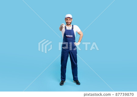 Full length happy courier in overalls, cap and protective gloves standing, pointing finger to camera, choosing you. Profession of service industry, loader or post delivery. indoor studio shot isolated 87739870
