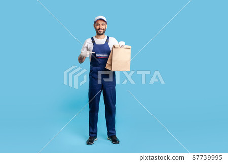 Full length portrait of courier in blue uniform pointing at groceries bag and looking at camera with toothy smile, carrying parcel with goods ordered online. Professional delivery service. studio shot 87739995