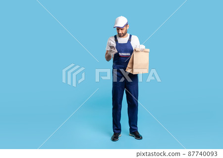 Full length responsible expert courier in blue uniform holding groceries bag and chatting with client on cell phone, reading message, using mobile app to accept online order. studio shot isolated Full length responsible expert courier in blue uniform holding groceries bag and chatting with client on cell phone, reading message, using mobile app to accept online order. studio shot isolated 87740093