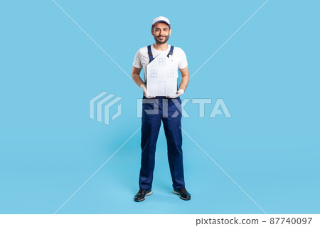 Full length handyman, building worker in uniform holding paper house. Mover guy offering help in relocation, home repair services and maintenance. indoor studio shot isolated on blue background Full length handyman, building worker in uniform holding paper house. Mover guy offering help in relocation, home repair services and maintenance. indoor studio shot isolated on blue background 87740097