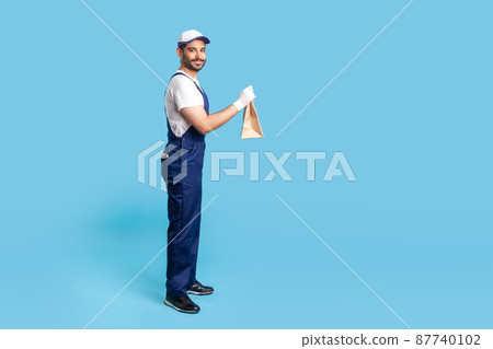 Side view, happy courier in blue uniform holding groceries bag and smiling to camera, carrying parcel with goods ordered online. Professional delivery service, food purchase. studio shot isolated 87740102
