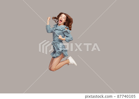 Delighted enthusiastic girl ruffle dress flying mid-air with raised fists shouting for joy, jumping trampoline looking at camera extremely happy, celebrating success. studio shot isolated on gray 87740105