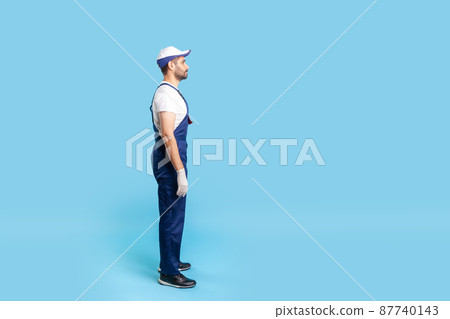 Full length profile of handyman in uniform standing calmly, looking serious at copy space, waiting order. Profession of service industry, house repair. indoor studio shot isolated on blue background 87740143