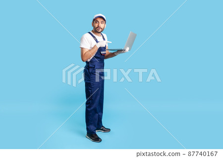 Full length amazed workman in uniform pointing at laptop and looking with shocked expression, surprised by message on computer, repair service order. indoor studio shot isolated on blue background 87740167