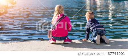 Back view two cute little curious sibling kids sitting on concrete embankment pier near sea lake bank and watching fish ship yacht boat marina. Children enjoy have fun playing outdoors at summer day Back view two cute little curious sibling kids sitting on concrete embankment pier near sea lake bank and watching fish ship yacht boat marina. Children enjoy have fun playing outdoors at summer day 87741901