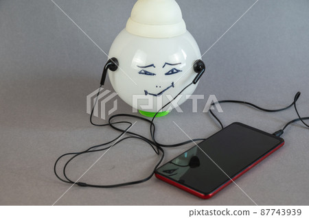 A large white electric lamp with a man's face painted on it. A smartphone and wired headphones. Composition against a gray background. 87743939