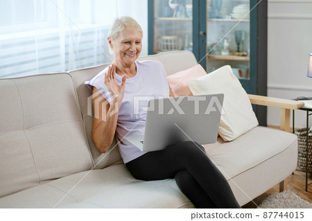 elderly woman sitting in a yoga pose in front of an open laptop. 87744015